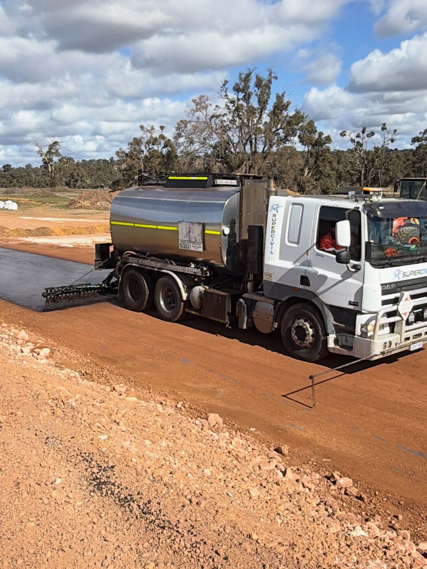 SuperCivil-branded bitumen sprayer truck applying spray seal to a rural red-dirt road in regional Western Australia