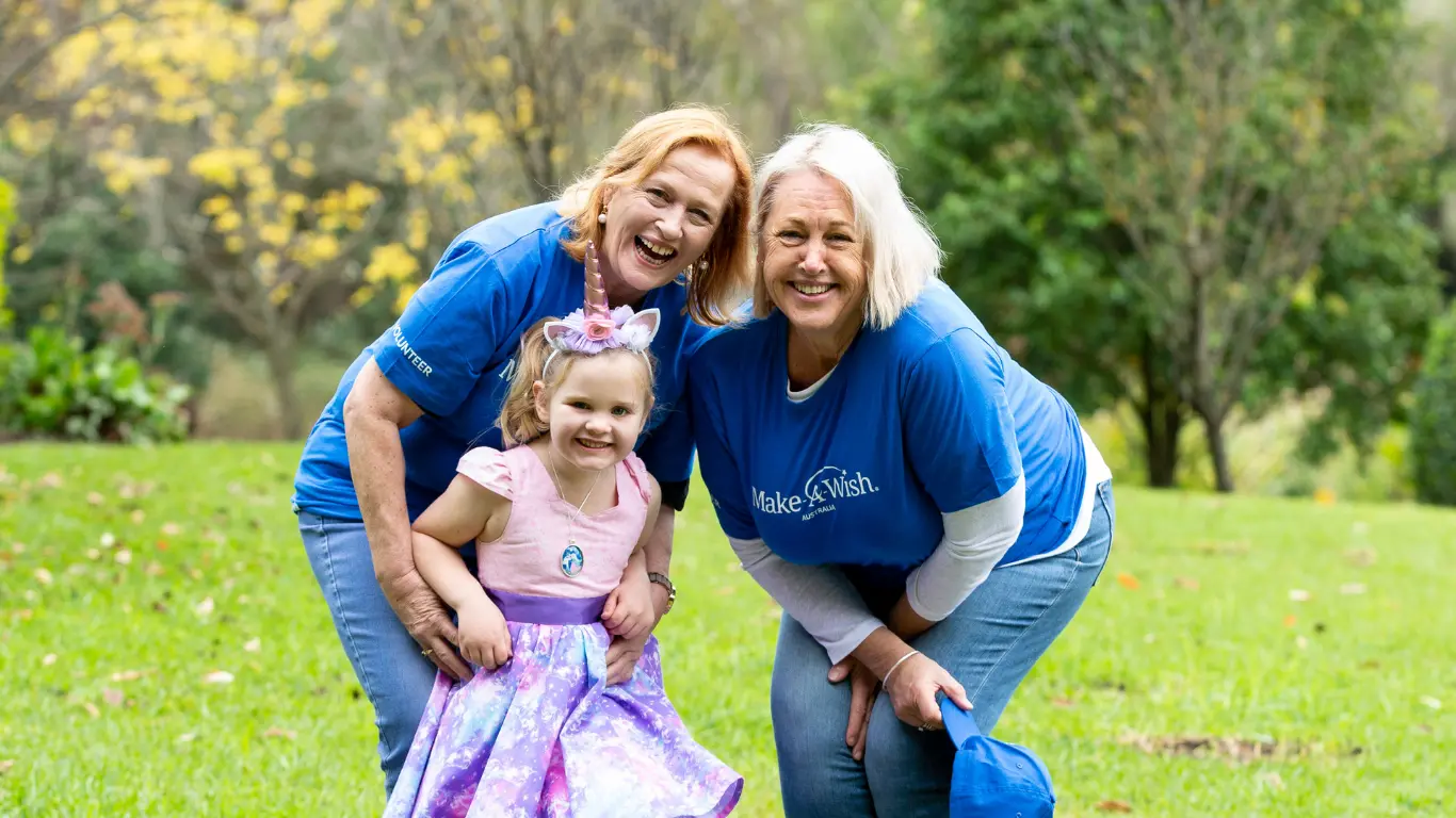 Make-A-Wish Australia volunteers smiling beside a young girl in a unicorn headband on a grassy lawn