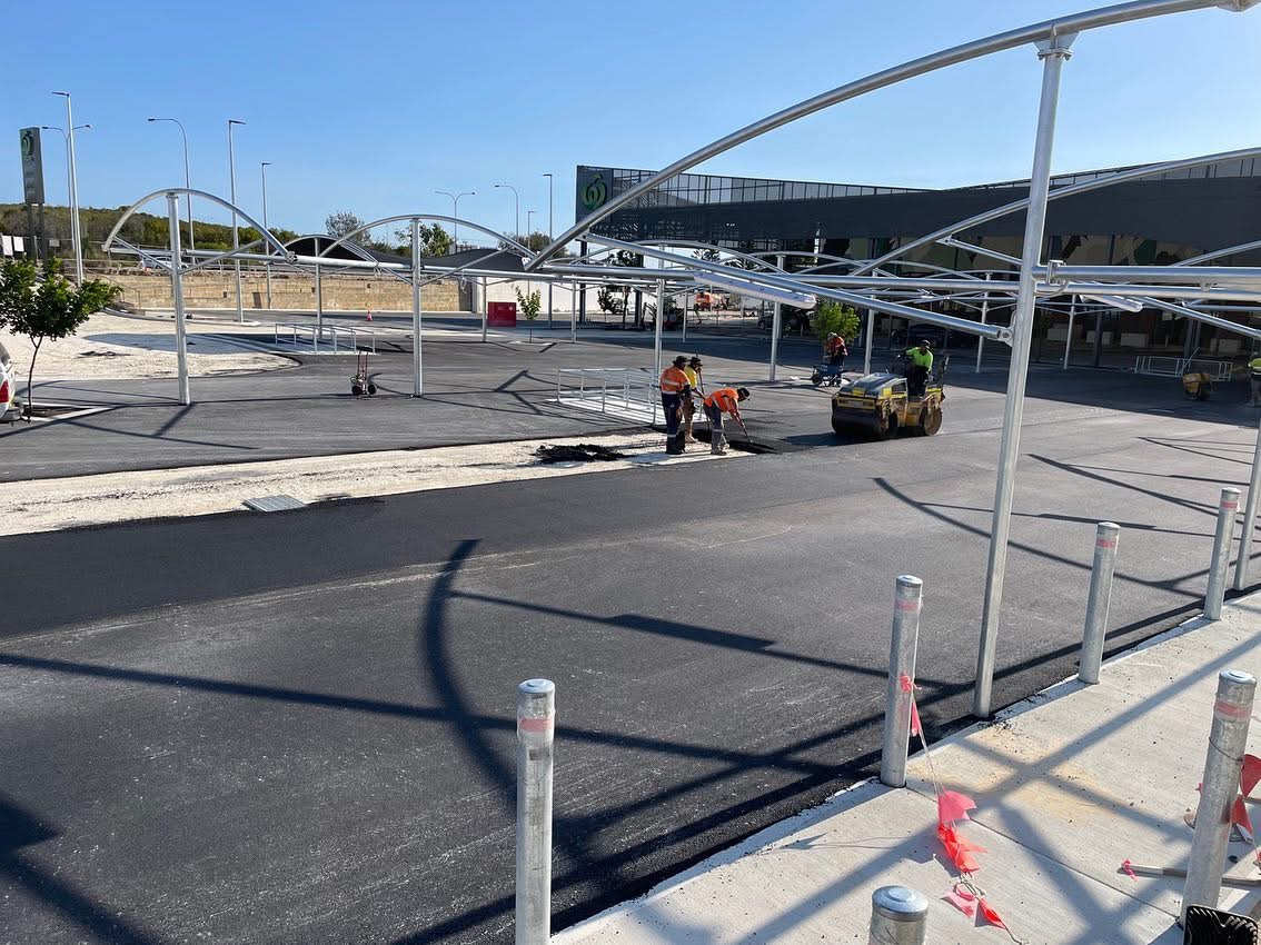 SuperCivil crew laying and compacting asphalt in a Woolworths shopping centre car park with curved shade sail canopies