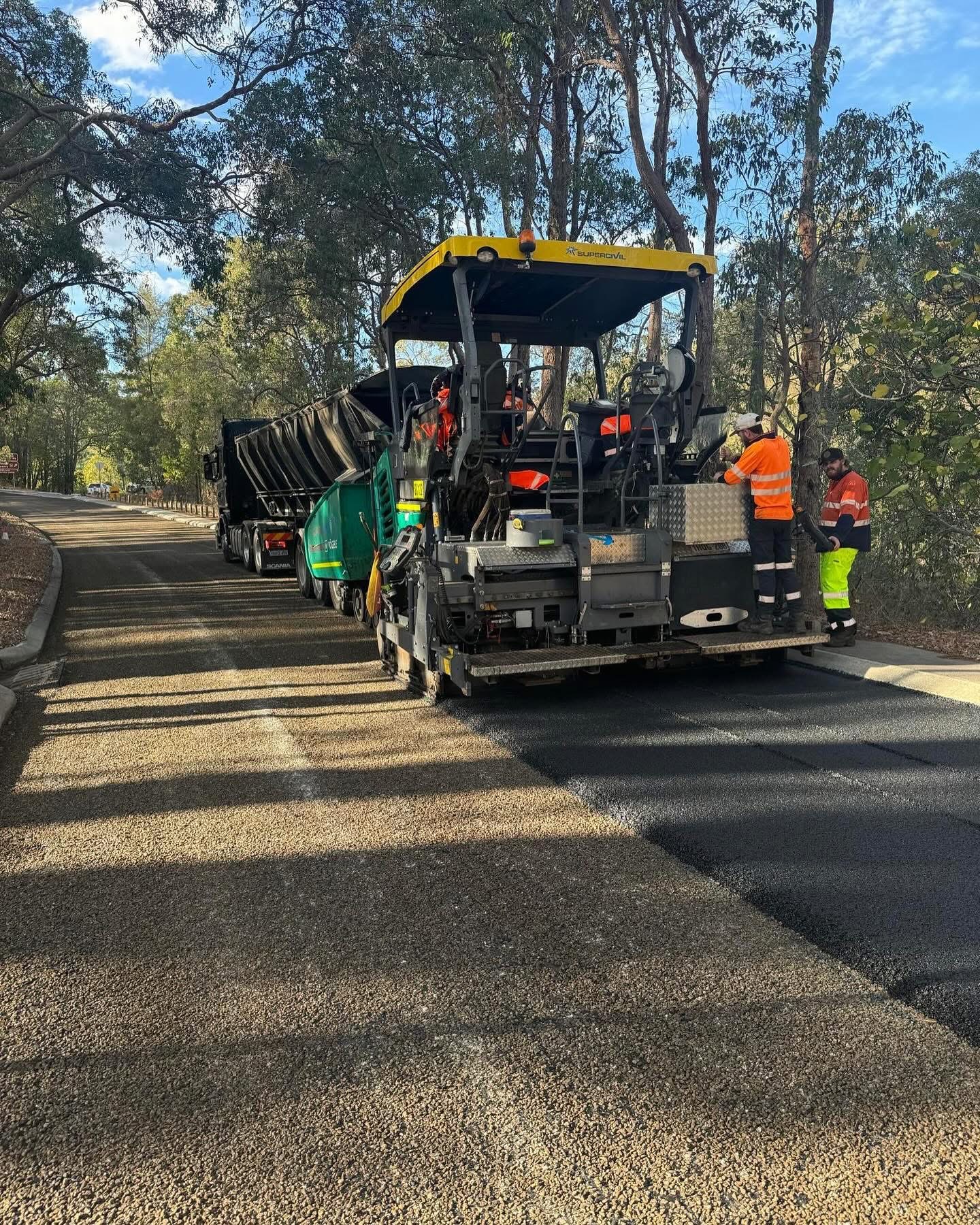 SuperCivil branded asphalt paver laying fresh asphalt on a tree-lined regional road with a Scania tipper truck feeding hot mix and hi-vis crew working