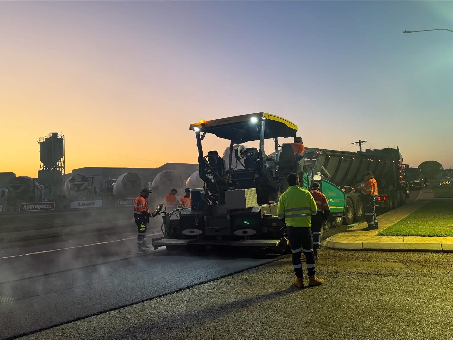 SuperCivil crew resurfacing a road at dawn with a Vögele paver laying fresh asphalt beside a tipper truck and aggregate supply yard