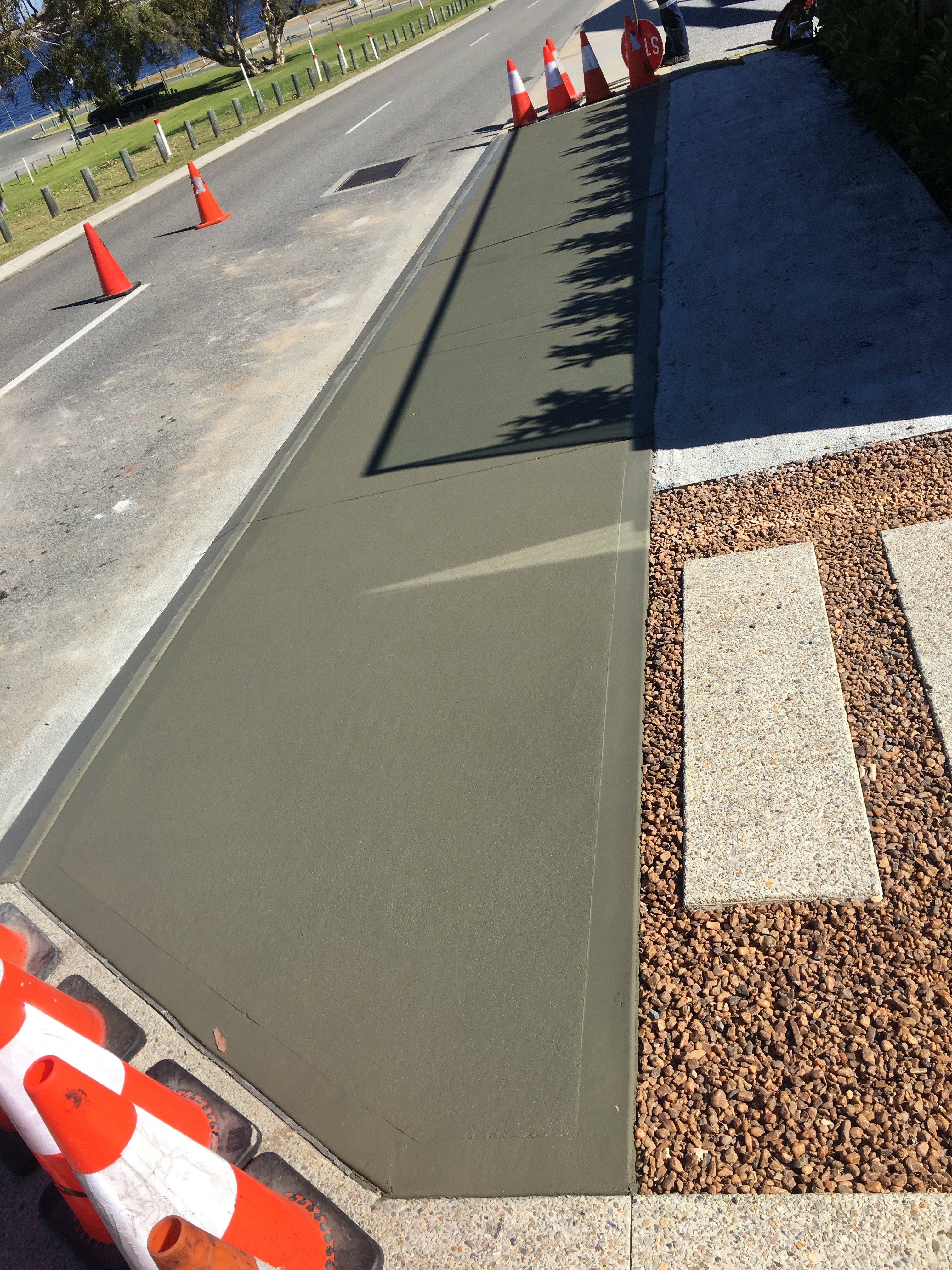 Freshly poured and finished roadside concrete footpath cordoned off with traffic cones, bordered by decorative gravel and pavers on a Western Australian project