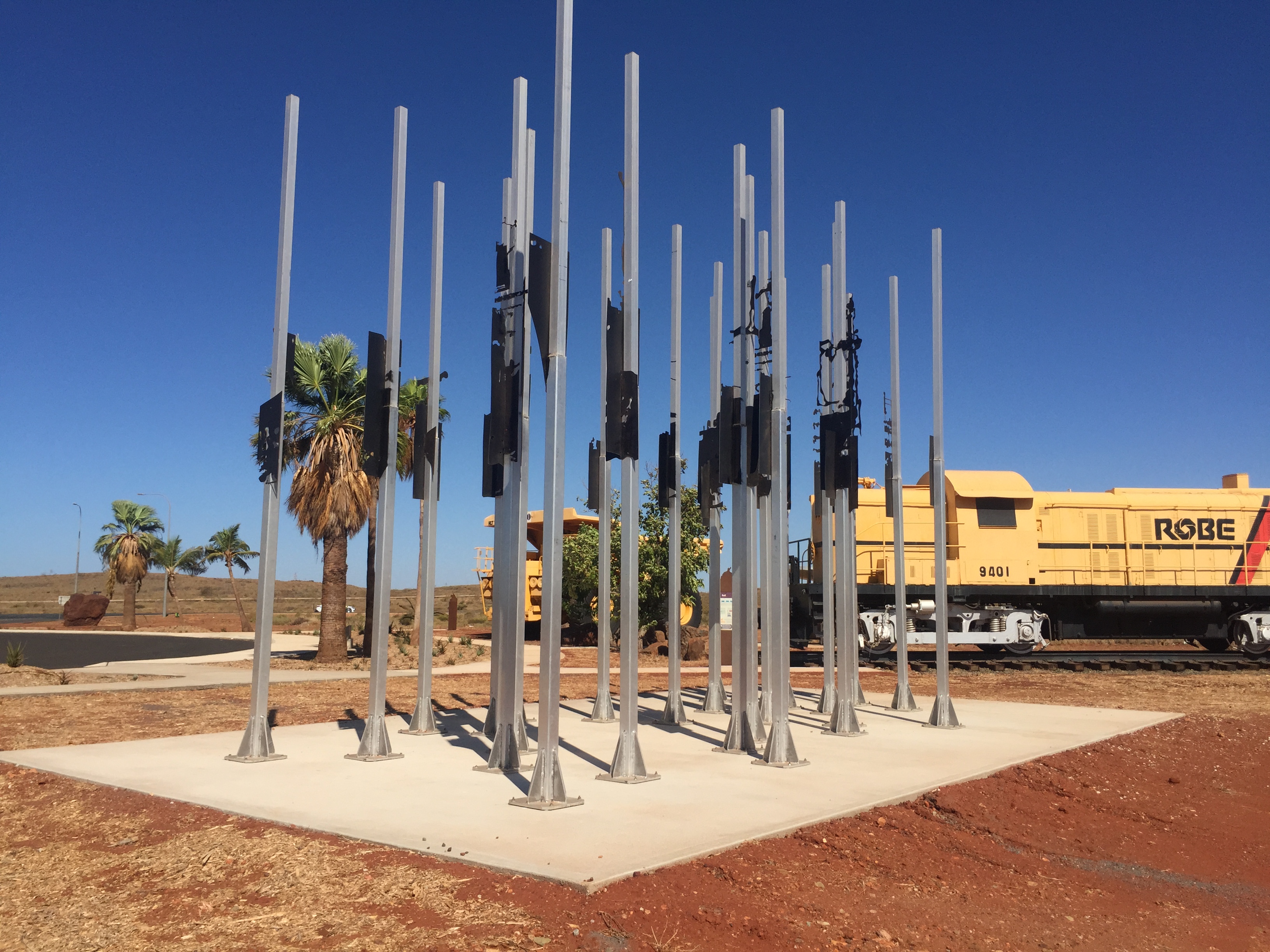 Public art installation of steel poles mounted on a poured concrete pad beside a heritage locomotive display in a regional Western Australian town plaza built by SuperCivil