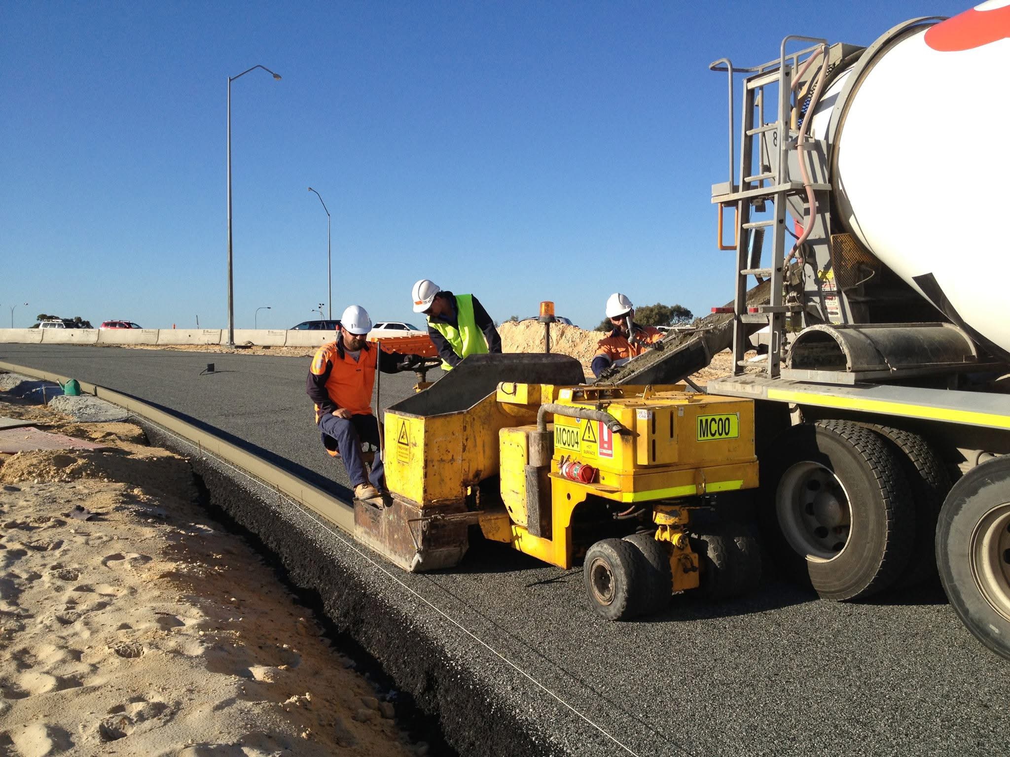 SuperCivil crew operating a slipform kerbing machine fed by a concrete agitator truck, extruding a fresh continuous concrete kerb along a newly paved Western Australian road