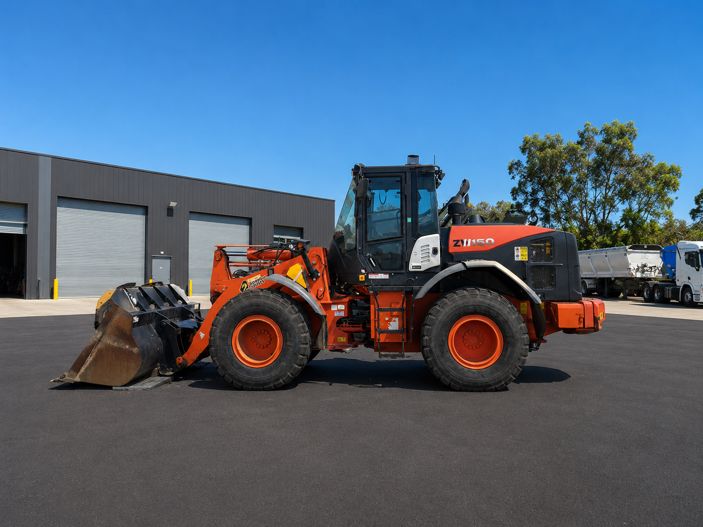 Orange Hitachi ZW150 wheel loader with a raised front bucket parked on asphalt outside a SuperCivil depot in Western Australia