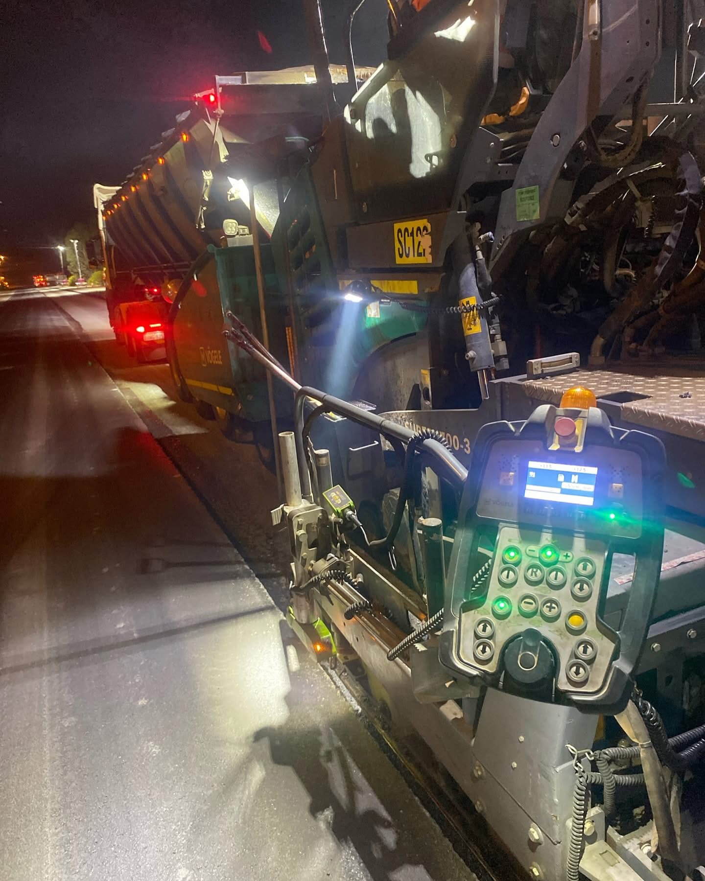 SuperCivil Vogele asphalt paver working under floodlights during night road construction works, with its illuminated control console in the foreground and a material feed truck ahead