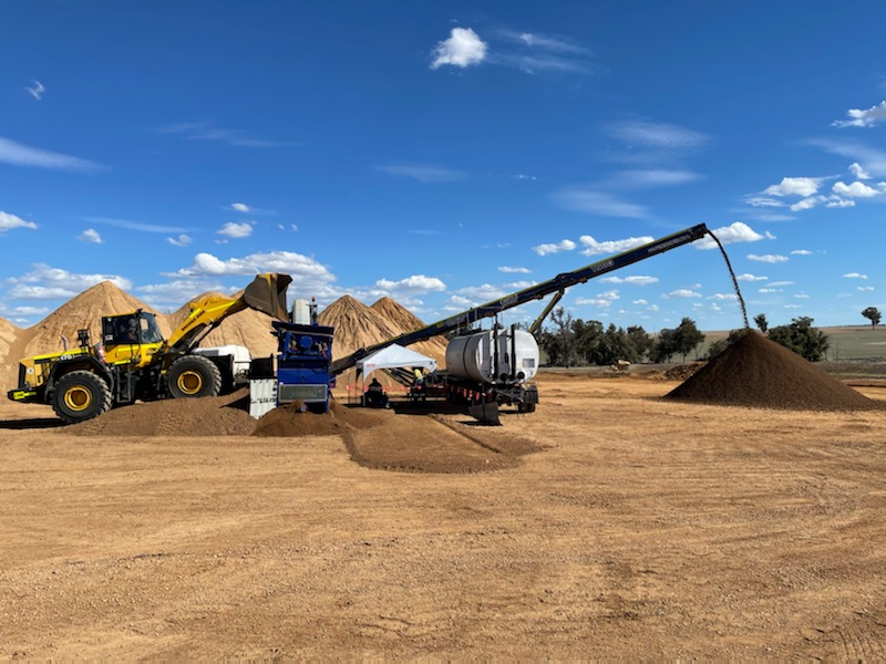 Blue SuperCivil pugmill plant blending stabilised material on a rural Western Australian site, with a wheel loader feeding stockpiled material and the conveyor discharging into a large stockpile under a blue sky