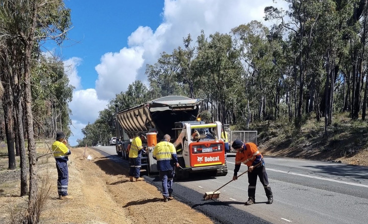 Hi-vis SuperCivil crew carrying out shoulder construction on a regional Western Australian highway with a Bobcat skid steer and tipper truck laying fresh asphalt while workers rake and sweep the new road shoulder