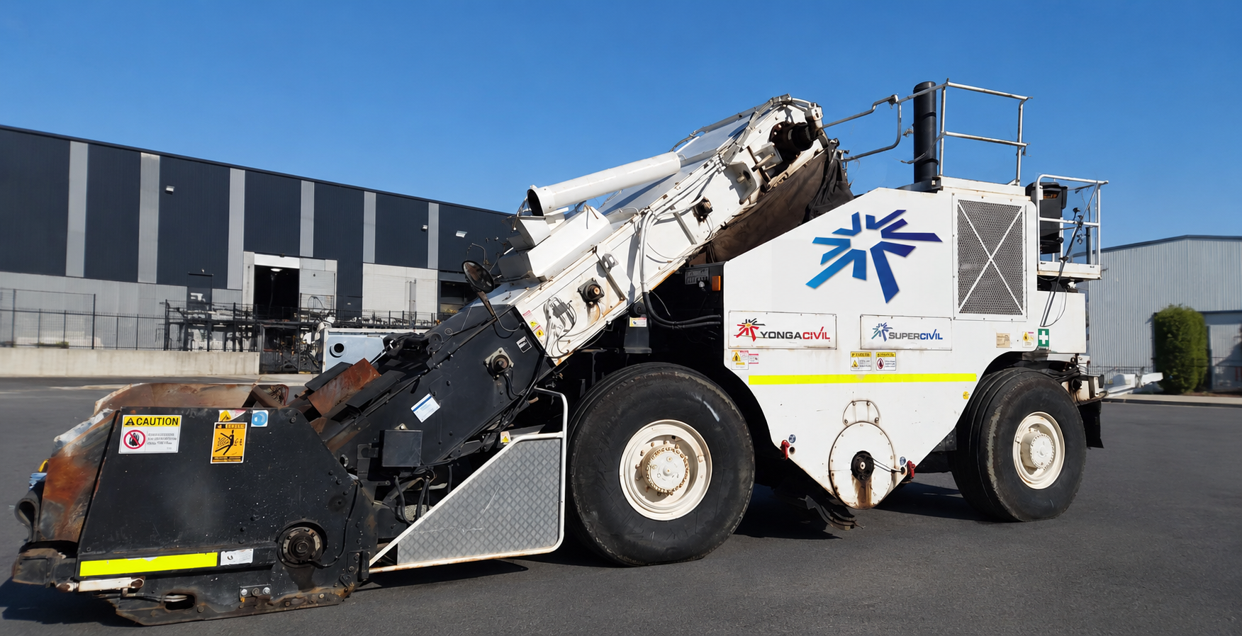 White SuperCivil shuttle buggy material transfer vehicle with raised conveyor arm, parked outside an industrial warehouse under a clear blue sky in Western Australia