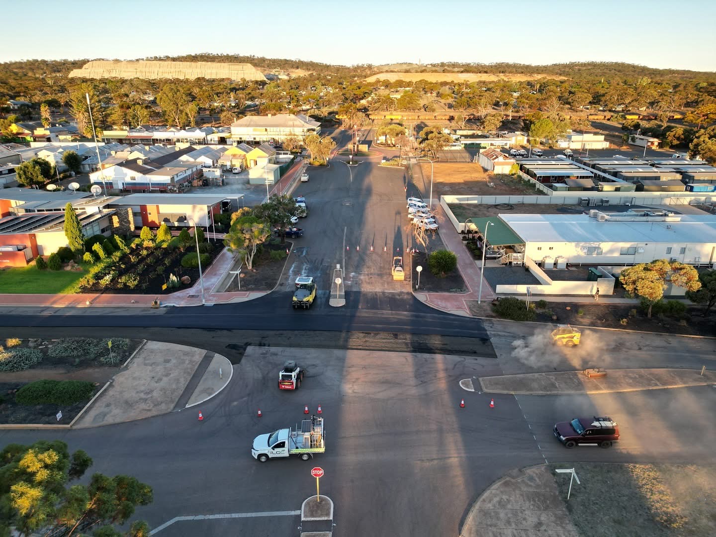 Aerial view of a regional Western Australian town intersection being resurfaced with fresh asphalt at golden hour, with a roller, paver and SuperCivil traffic management vehicles closing the road