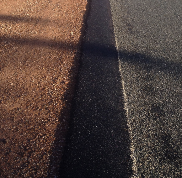 Close-up of a freshly sealed dark bitumen pavement edge strip between the red-dirt gravel shoulder and the existing asphalt road surface with a white edge line