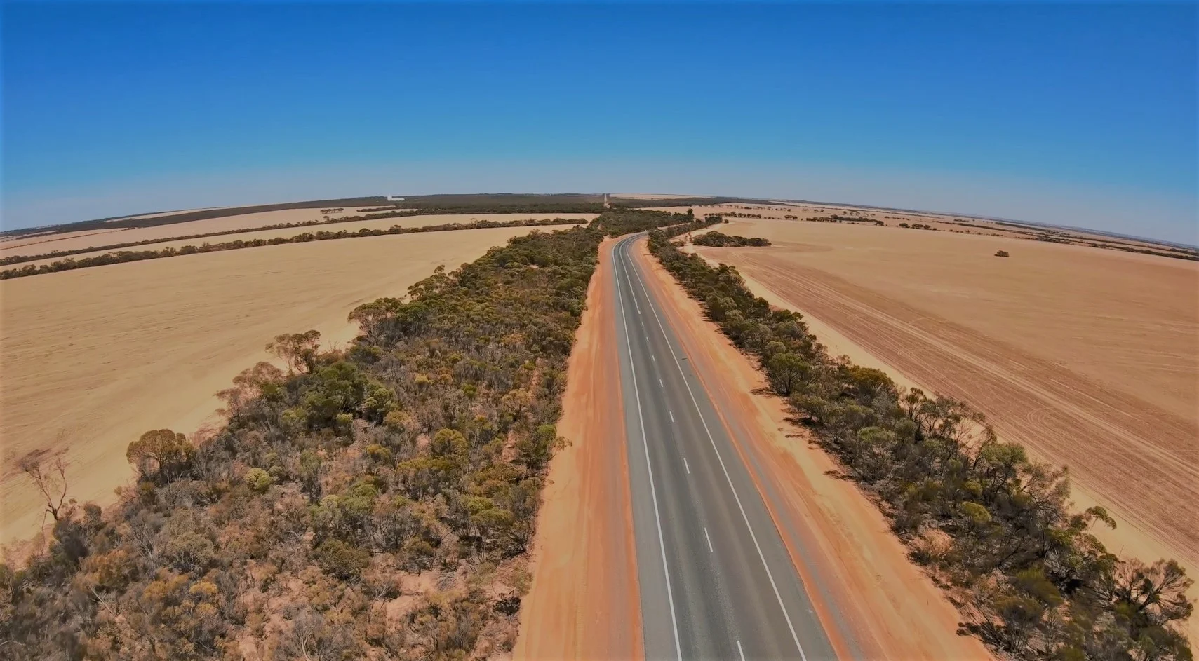 Aerial view of a straight sealed two-lane rural Western Australian highway running through golden cropping fields and red-dirt verges under a clear blue sky