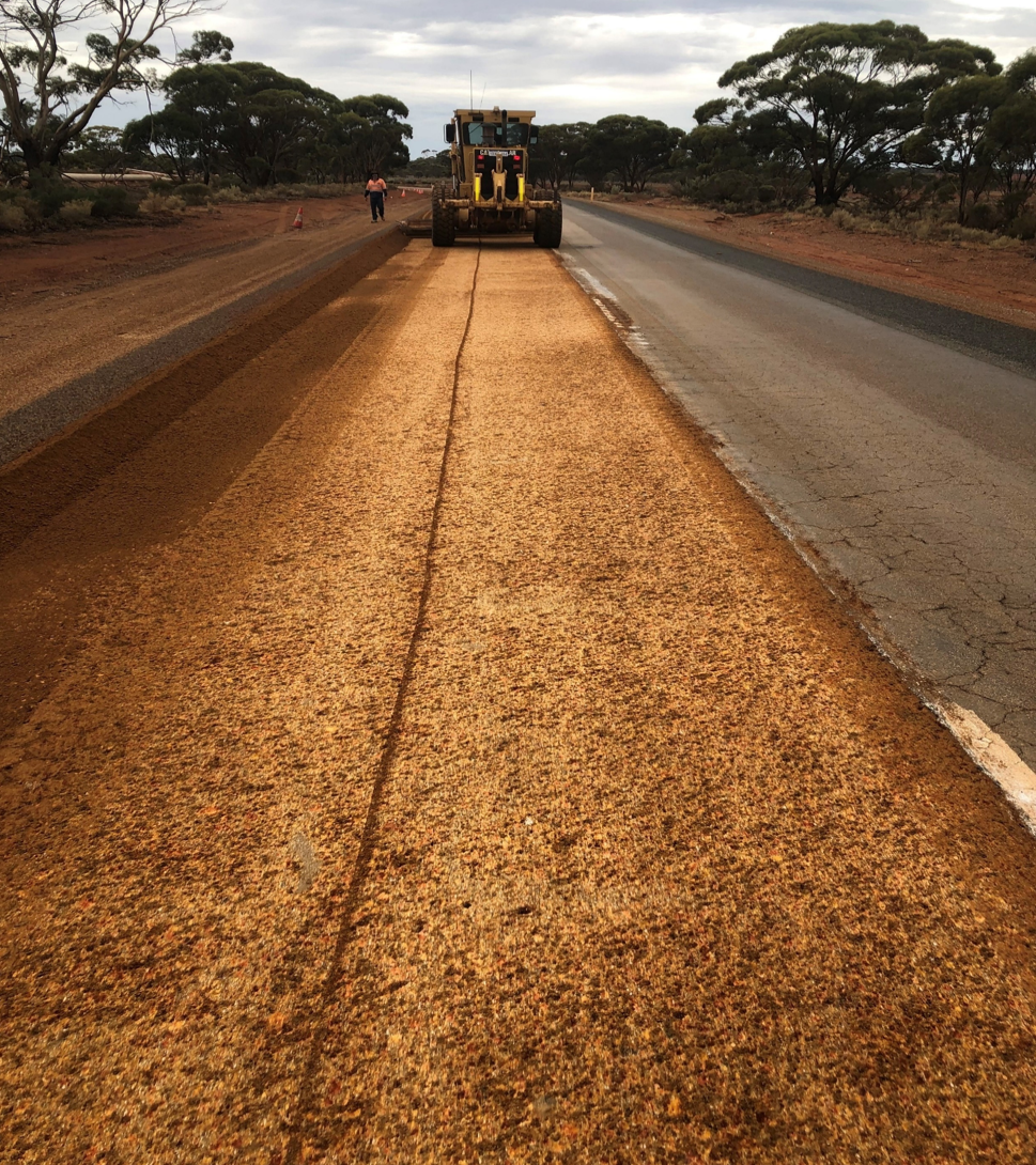 Caterpillar grader reshaping a freshly graded red-dirt road base alongside an existing sealed lane during road maintenance on a regional Western Australian highway