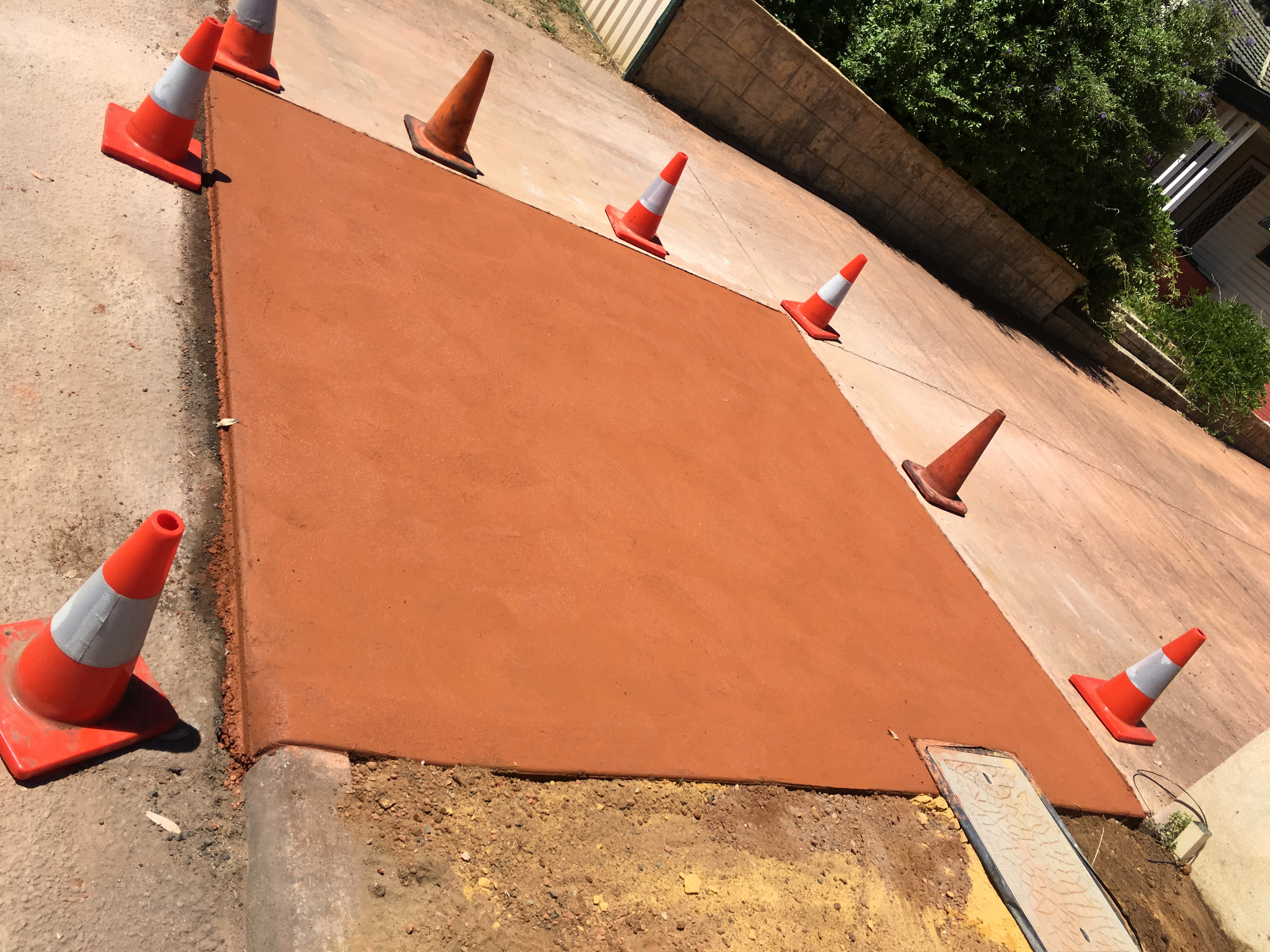 Freshly reinstated red-oxide concrete footpath crossover cordoned off with traffic cones alongside existing paving in a Western Australian suburban streetscape