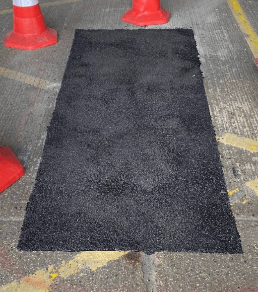 Freshly laid rectangular black asphalt pothole patch on a light grey road surface framed by red traffic cones