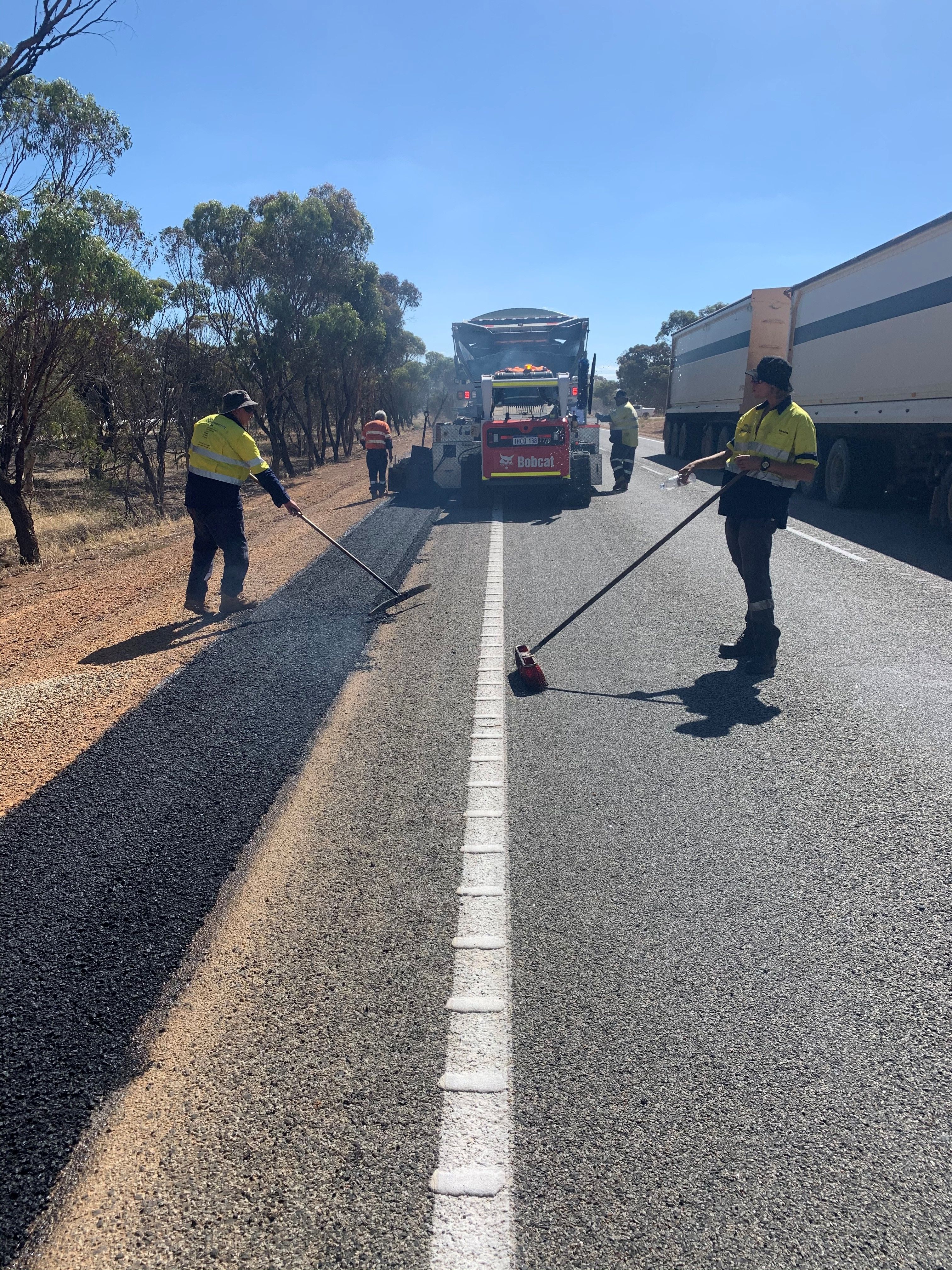 SuperCivil crew in hi-vis raking and spreading fresh asphalt to widen the shoulder of a regional Western Australian highway with a Bobcat skid-steer feeding material