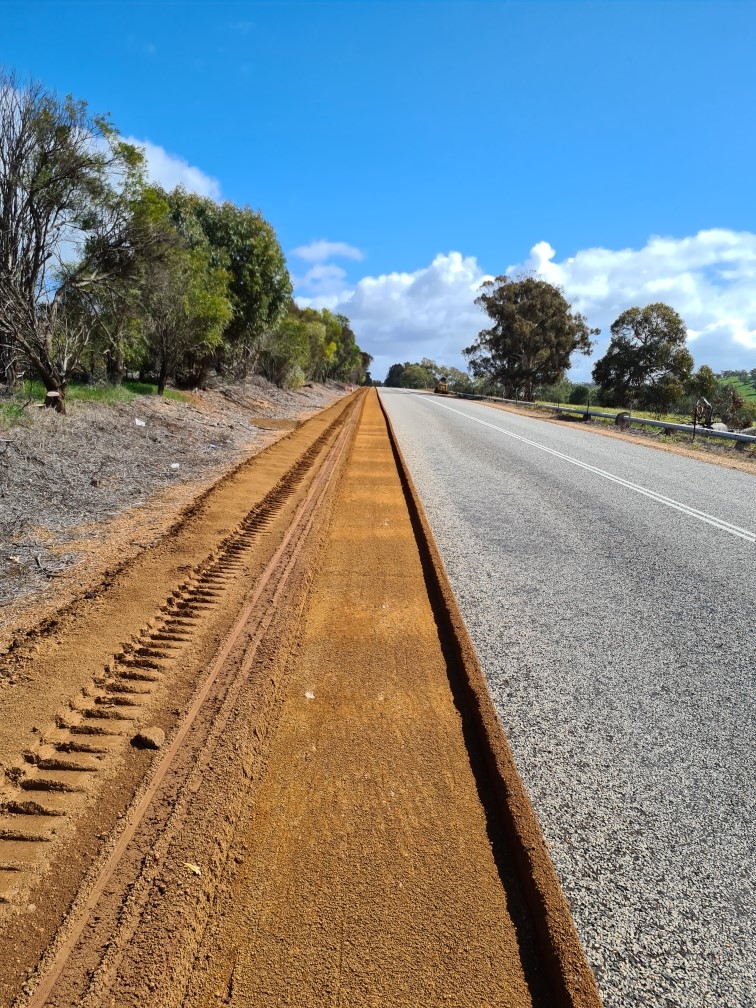 Freshly graded and compacted red-dirt road shoulder with grader track marks alongside a rural Western Australian highway