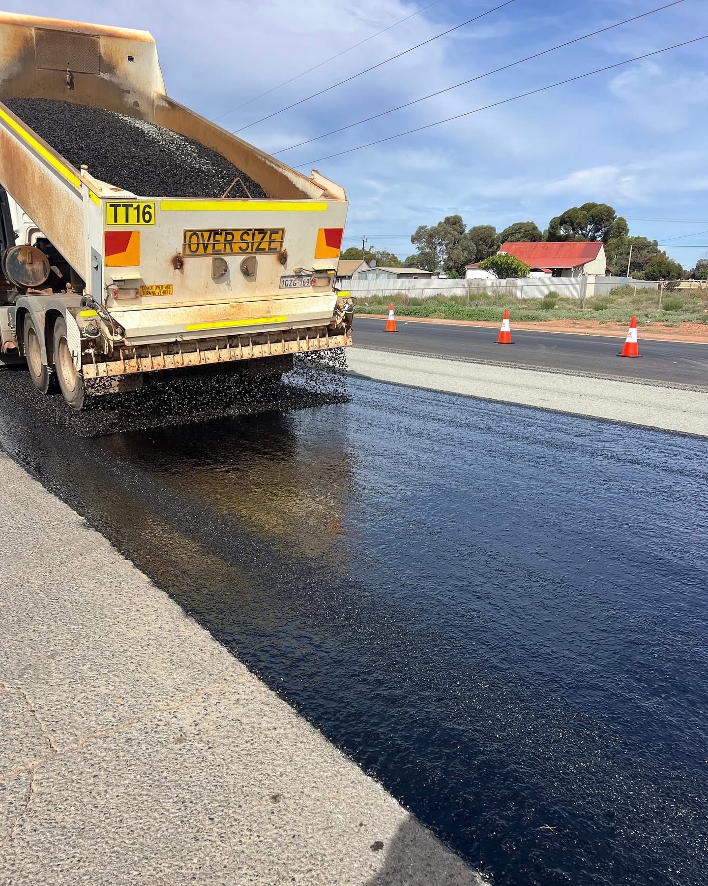 SuperCivil aggregate spreader truck applying freshly sprayed bituminous binder and aggregate across a Western Australian road during a spray sealing operation, with traffic cones and a glossy wet seal surface