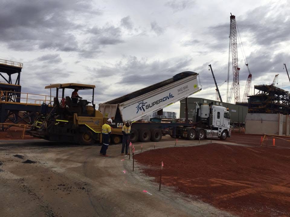 SuperCivil semi end tipper with its trailer raised discharging asphalt into a Caterpillar paver on a red-dirt mine site, with a hi-vis crew and cranes in the background