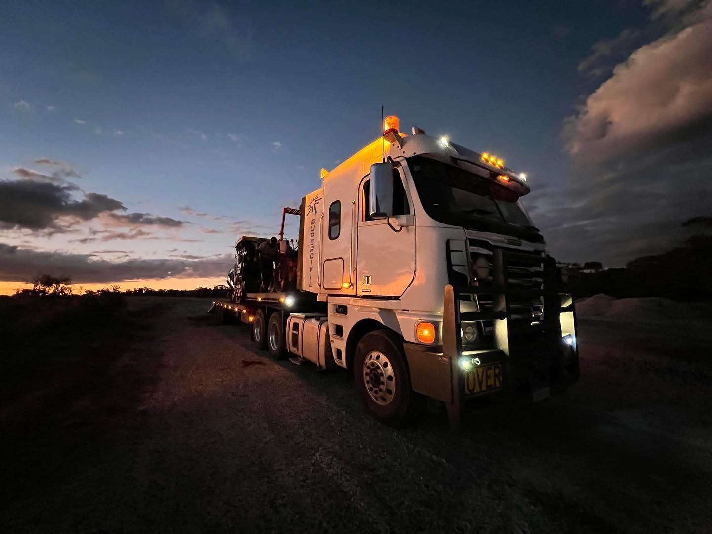 White SuperCivil prime mover hauling an oversized machine on a low-loader float, parked on a gravel pad at dusk with amber cab lights and a moody sunset sky over regional Western Australia