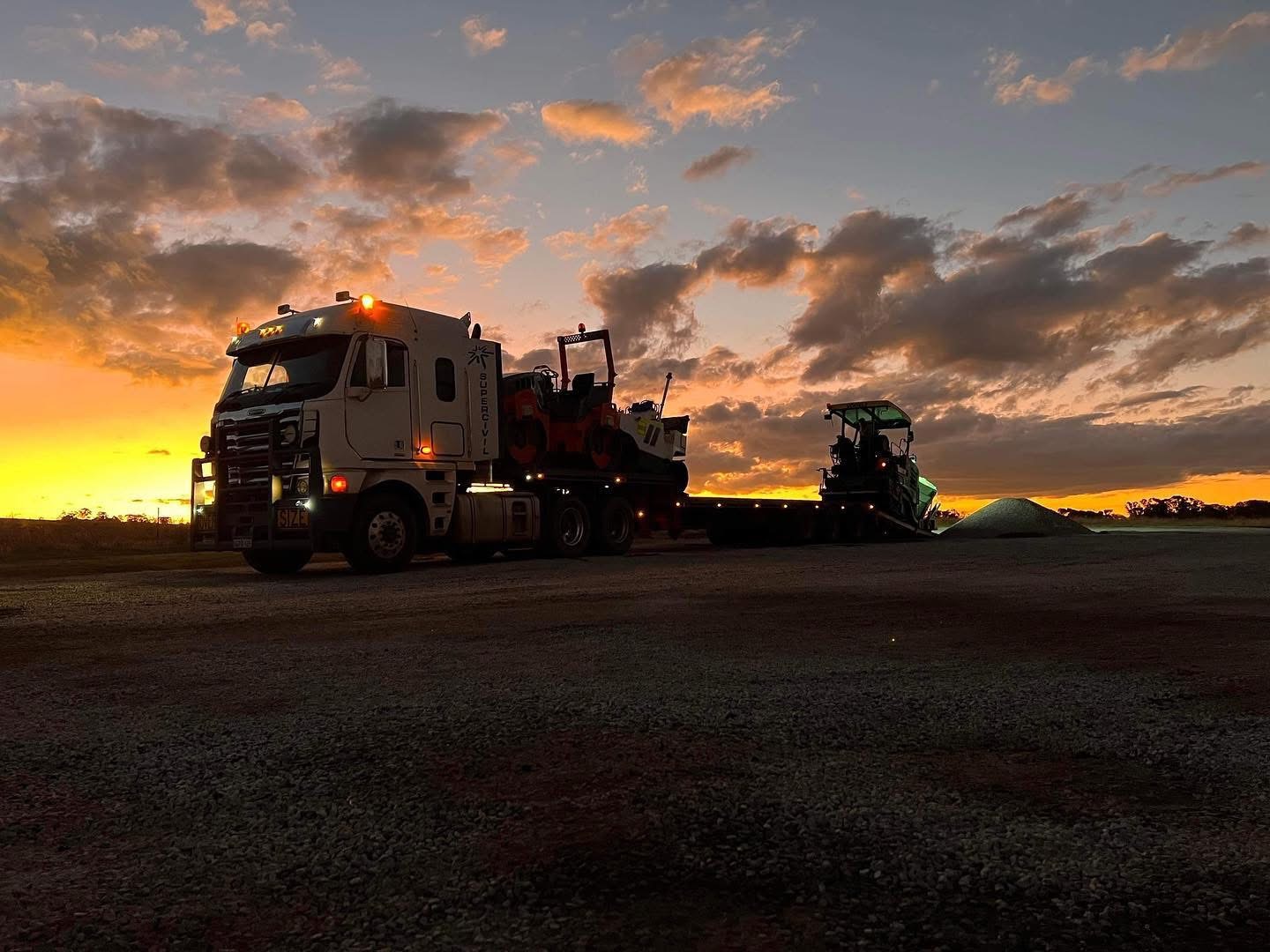 White SuperCivil prime mover with a low-loader float carrying a compaction roller and civil plant, parked on a gravel pad at sunset in regional Western Australia