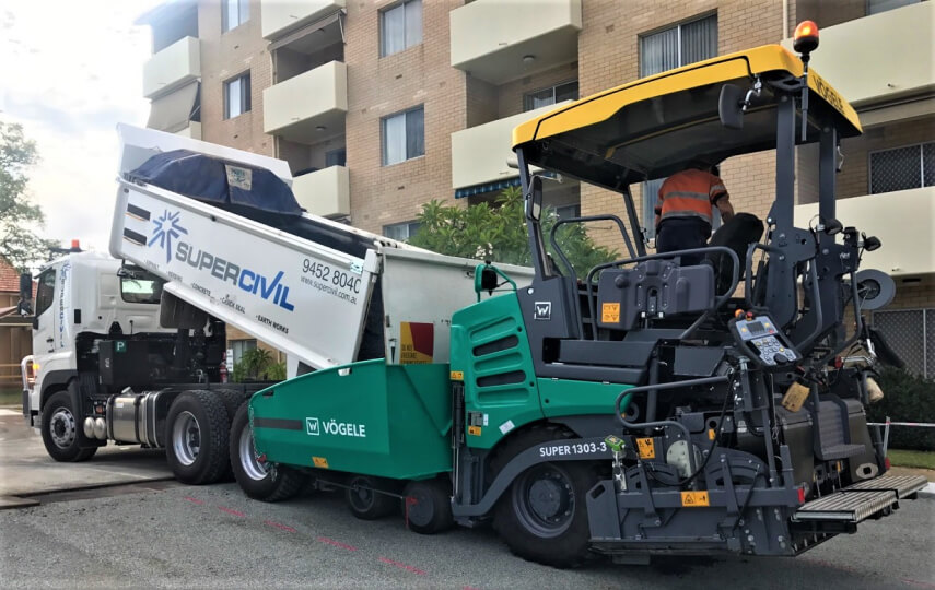 SuperCivil 6-wheel tipper with its body raised discharging asphalt into a green Vögele paver on a suburban street in front of a residential apartment block
