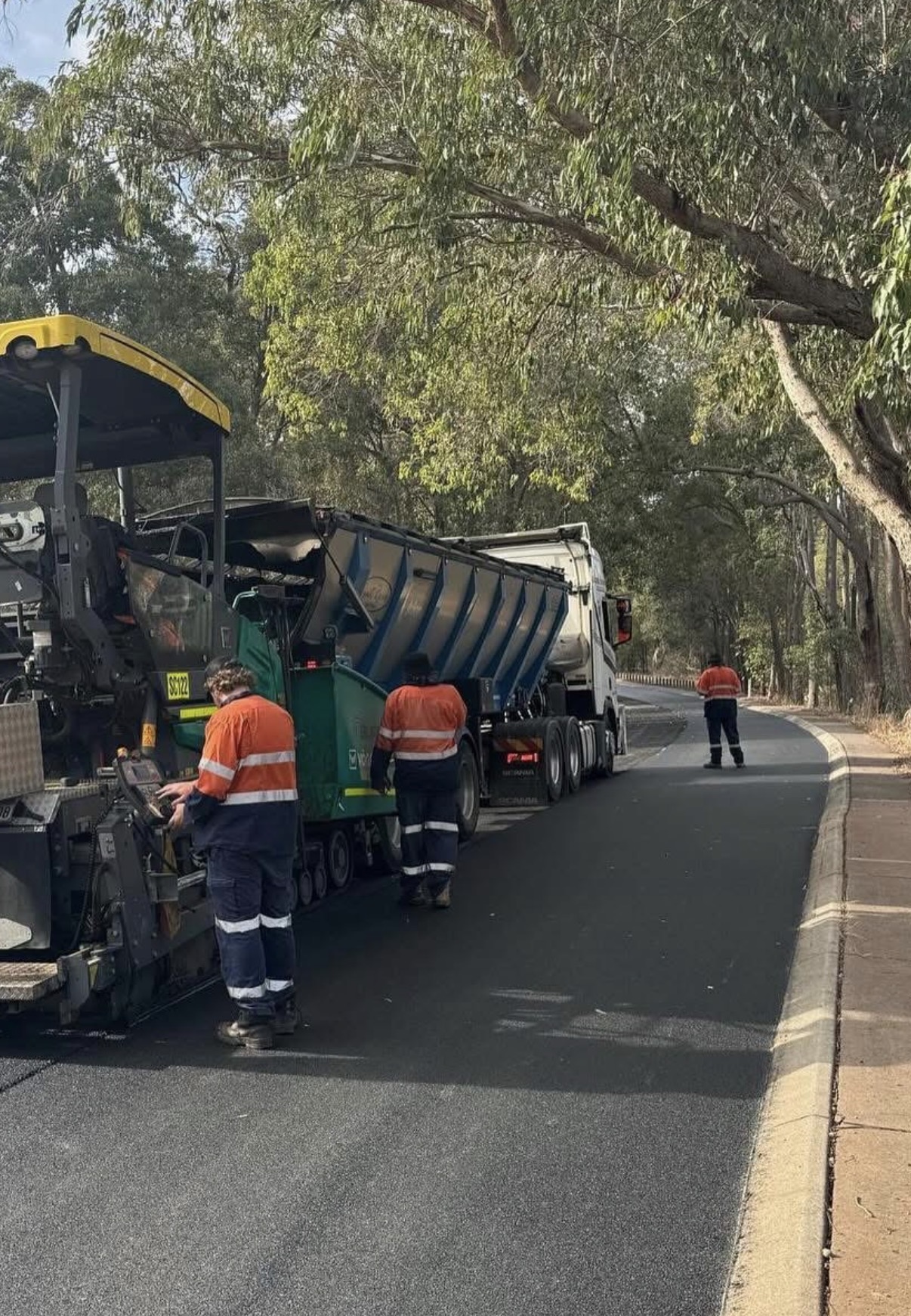 SuperCivil walking floor trailer feeding asphalt into a paver on a tree-lined suburban Perth road while a hi-vis crew lays a fresh asphalt mat